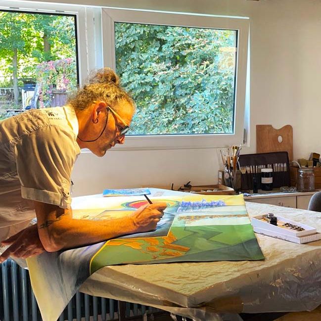 Der Künstler Erik Adam H Grösch in seinem Atelier bei Tuschearbeiten am Arbeitsplatz. Dahinter ein Blick aus dem Fenster ins Grüne.